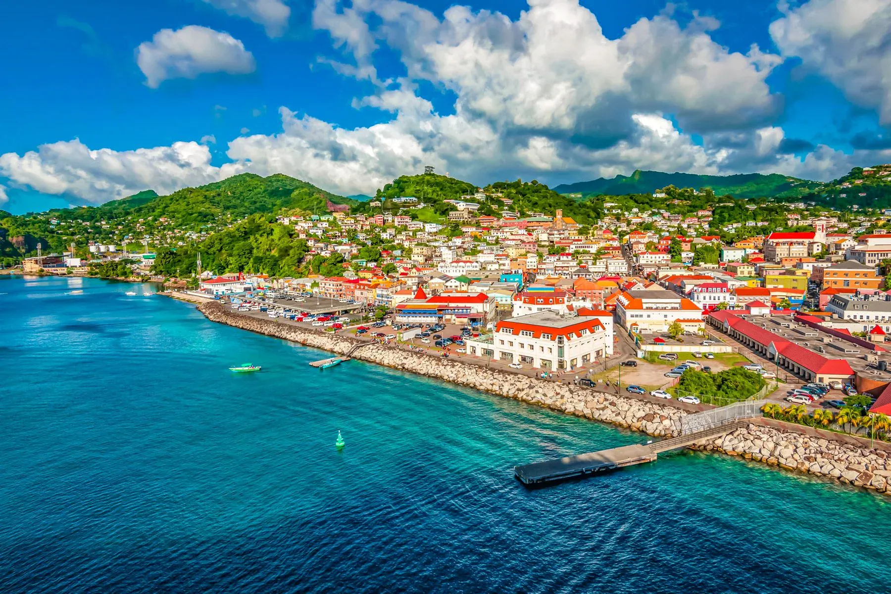 Aerial view of a coastal town with colorful buildings, green hills in the background, and a blue sea in the foreground under a partly cloudy sky.