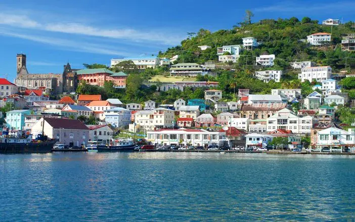 Colorful buildings and houses line a hillside above a waterfront in a coastal town with clear blue water and a bright blue sky—perfect scenery to explore with a convenient car rental in Grenada.