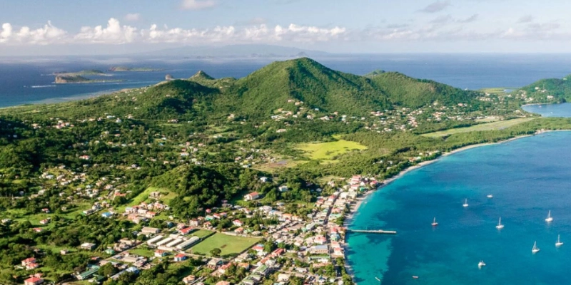 Aerial view of a coastal town with green hills, scattered houses, and a turquoise bay with boats anchored offshore—perfect for exploring with a car rental Grenada adventure.