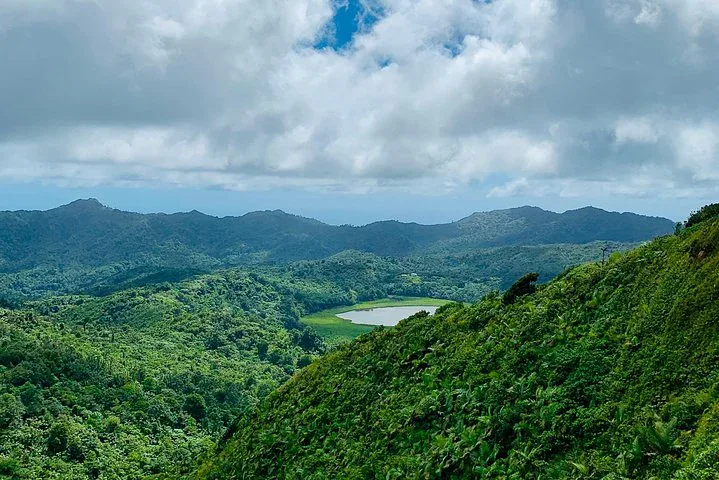 A lush green landscape with dense forest, rolling hills, a small lake in the center, and a partly cloudy sky overhead—perfect to explore with a car rental in Grenada.