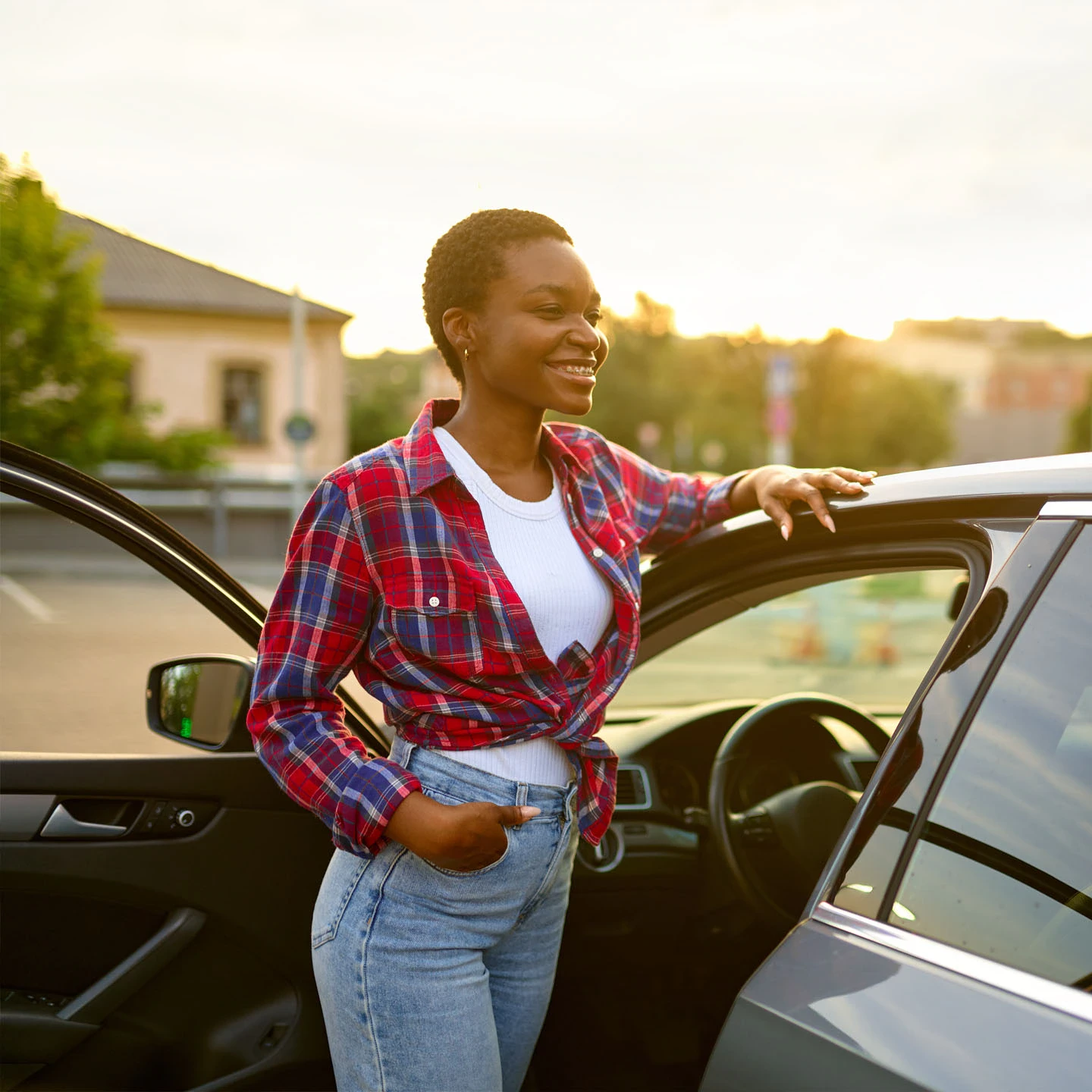 A person stands by an open car door, smiling, wearing a red plaid shirt and jeans in a parking lot during sunset, perhaps after visiting a car rental agency in Grenada.