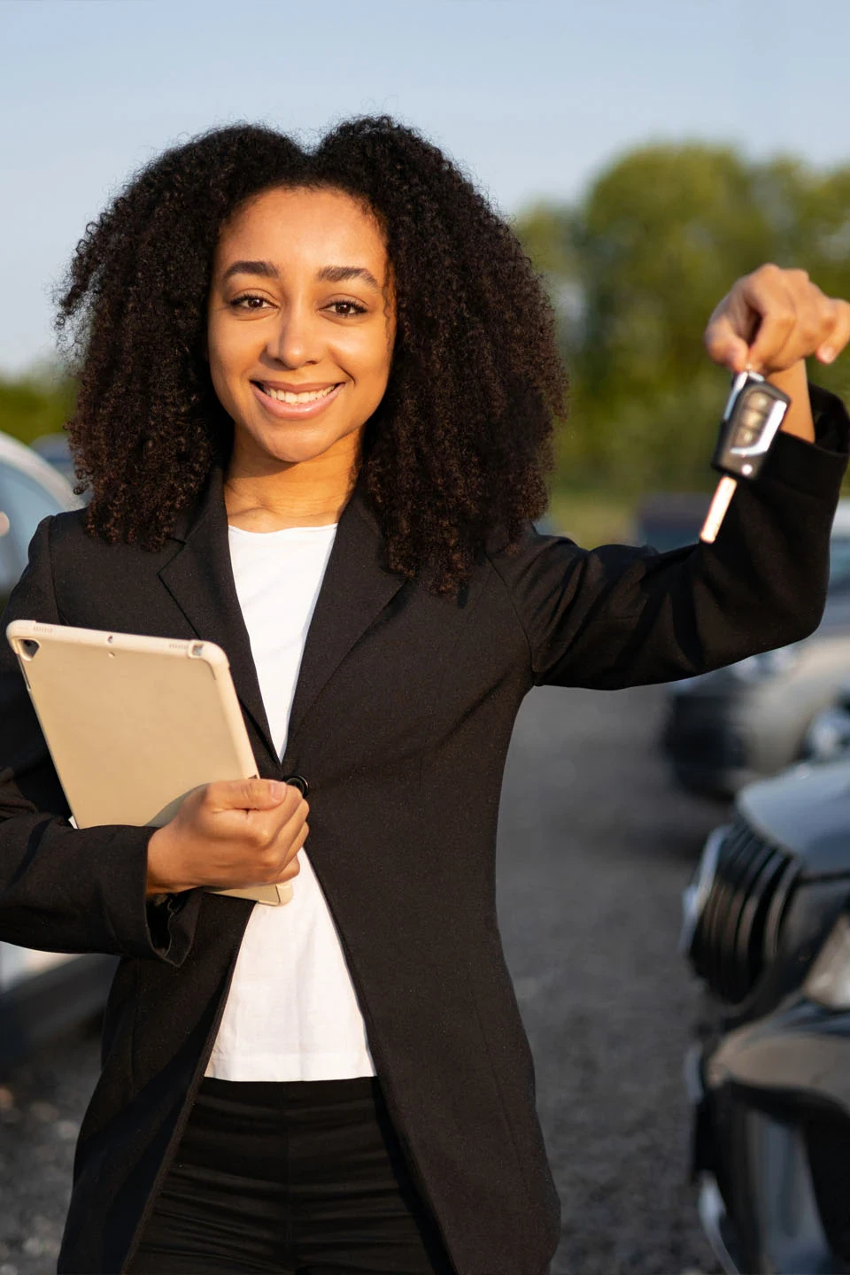 A woman in a black blazer stands outdoors holding a tablet and car keys, smiling at the camera, with cars visible in the background.